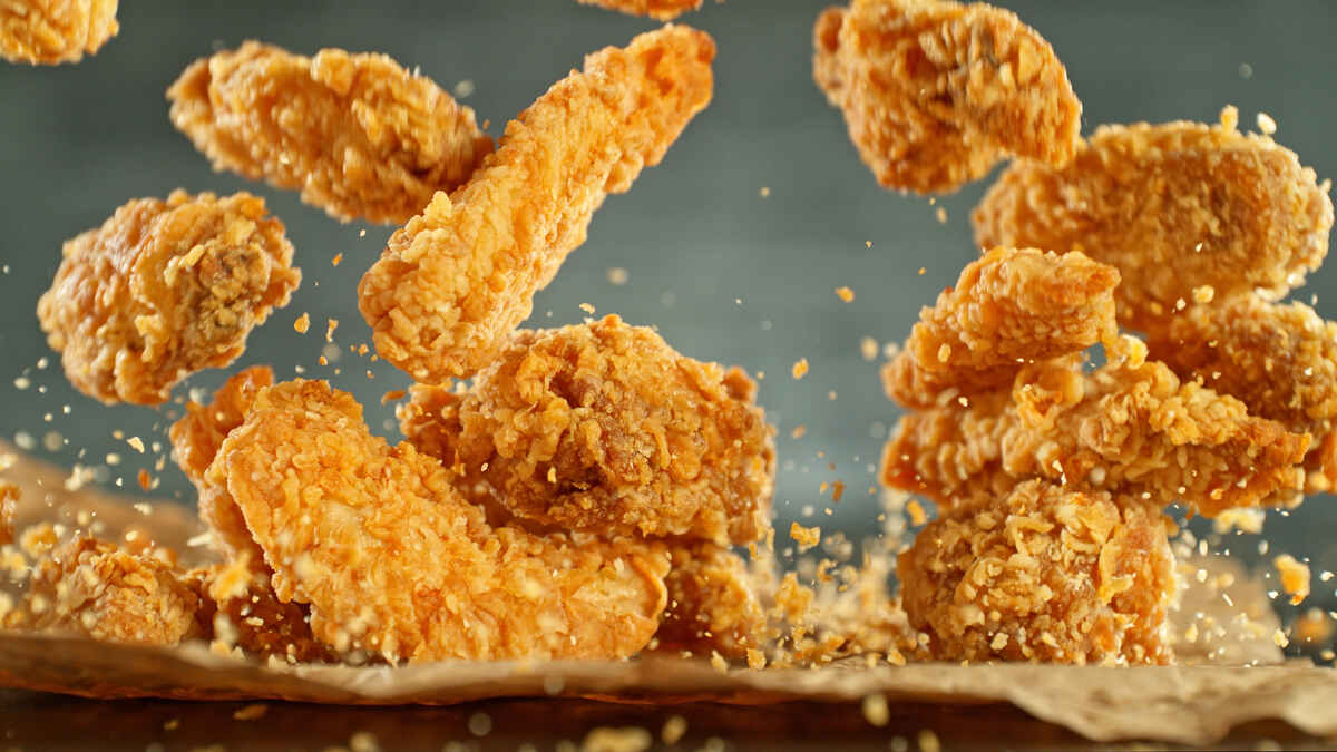 Several pieces of crispy fried chicken are shown mid-air above a brown paper surface, with crumbs scattered around.