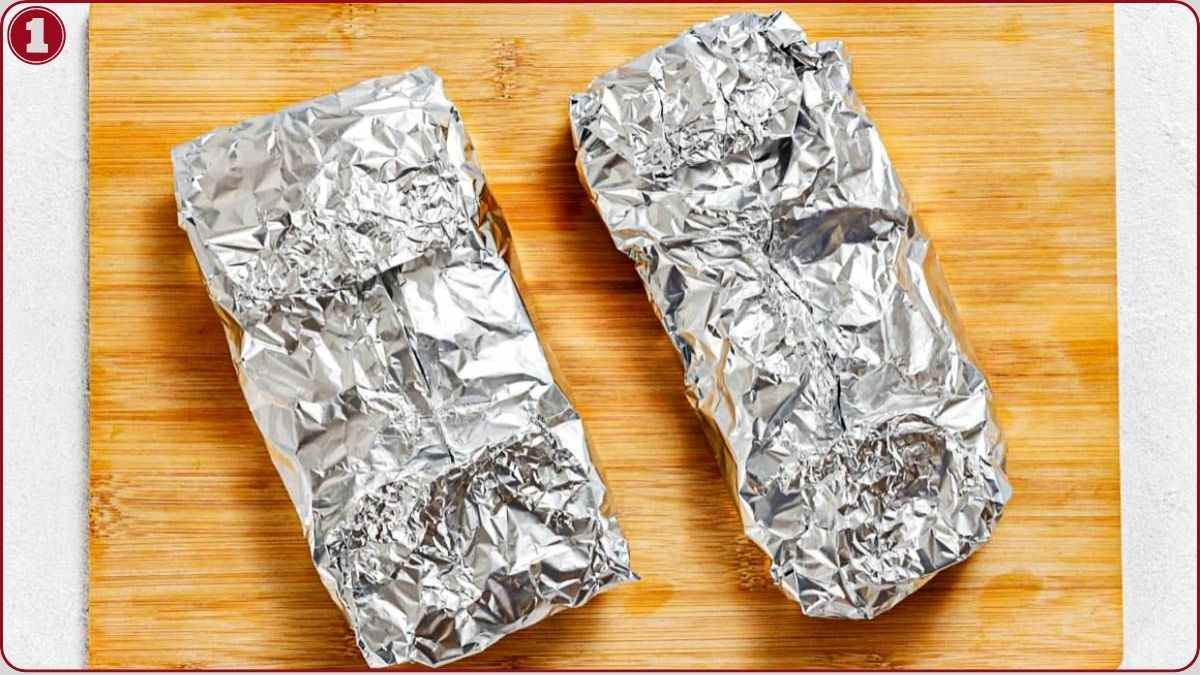 Two rectangular food items wrapped in aluminum foil are placed on a wooden cutting board.