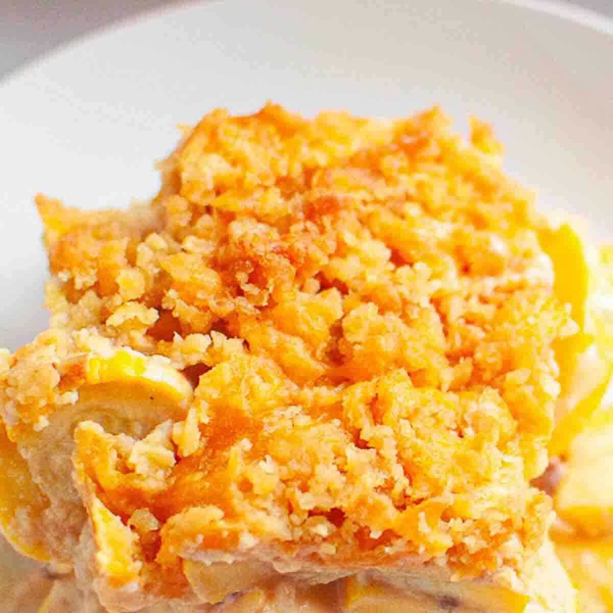 A close-up of a serving of baked casserole with a golden, crumbly topping, creamy filling, and visible yellow slices, presented on a white plate.