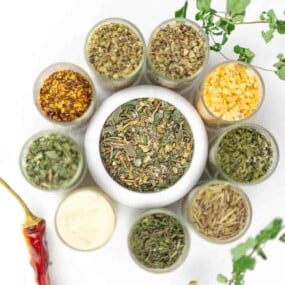 Top-down view of various dried herbs and spices in small glass containers arranged in a circle around a central bowl, with a dried chili pepper and green herbs nearby.