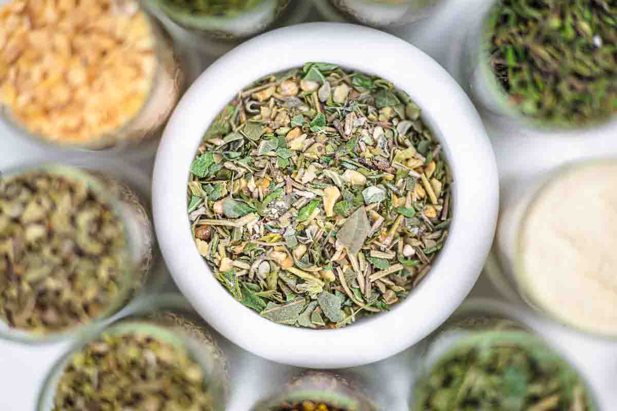 A white bowl filled with a mixed dried herb blend, surrounded by jars containing various other herbs and seasonings.
