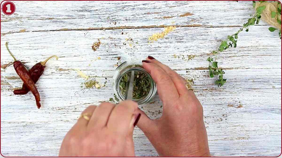 A person stirring dried herbs in a small glass jar on a white wooden surface, with chili peppers and fresh herbs nearby.