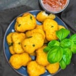 A plate of breaded, golden-brown nuggets garnished with fresh basil, served with a cup of marinara sauce.