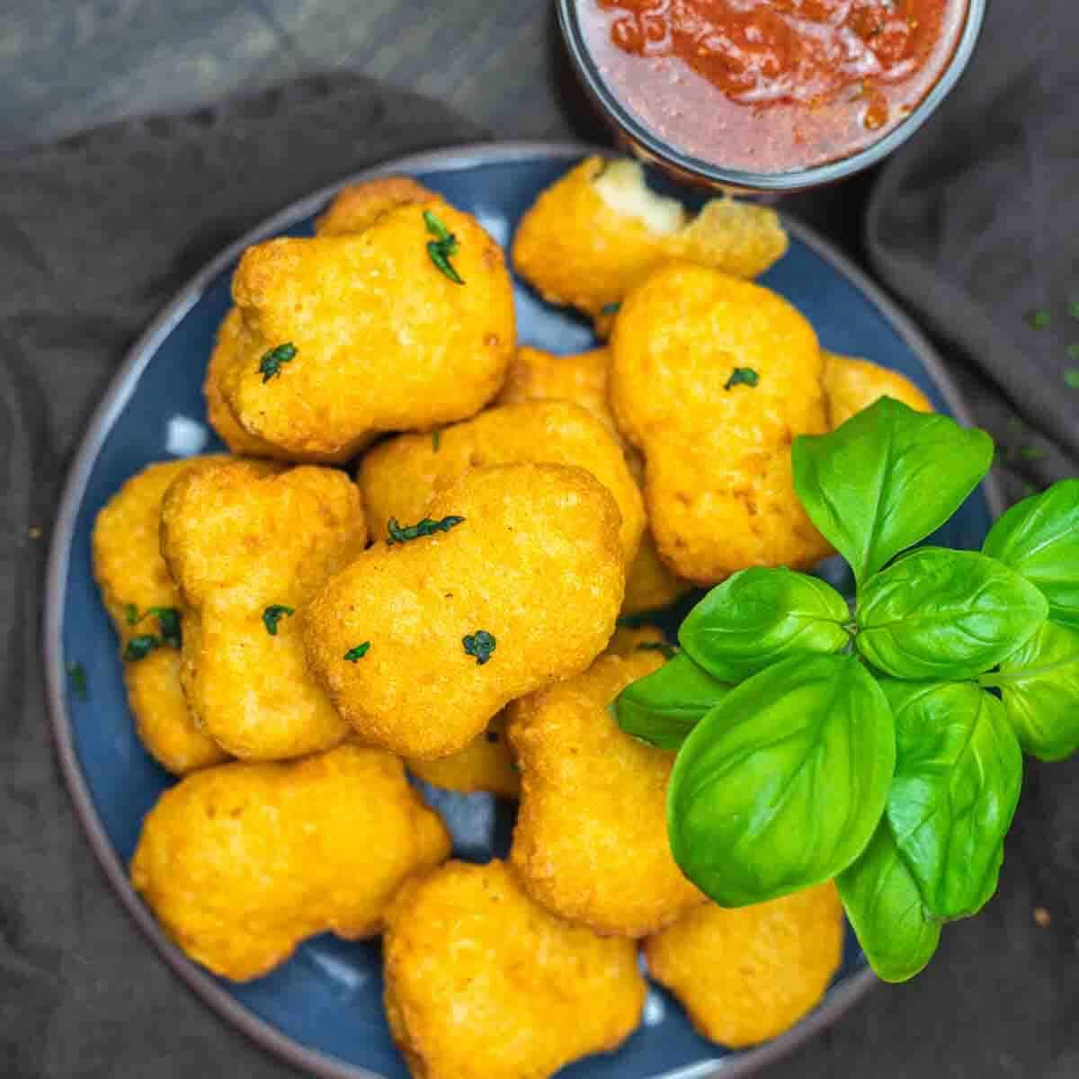 A plate of breaded, golden-brown nuggets garnished with fresh basil, served with a cup of marinara sauce.