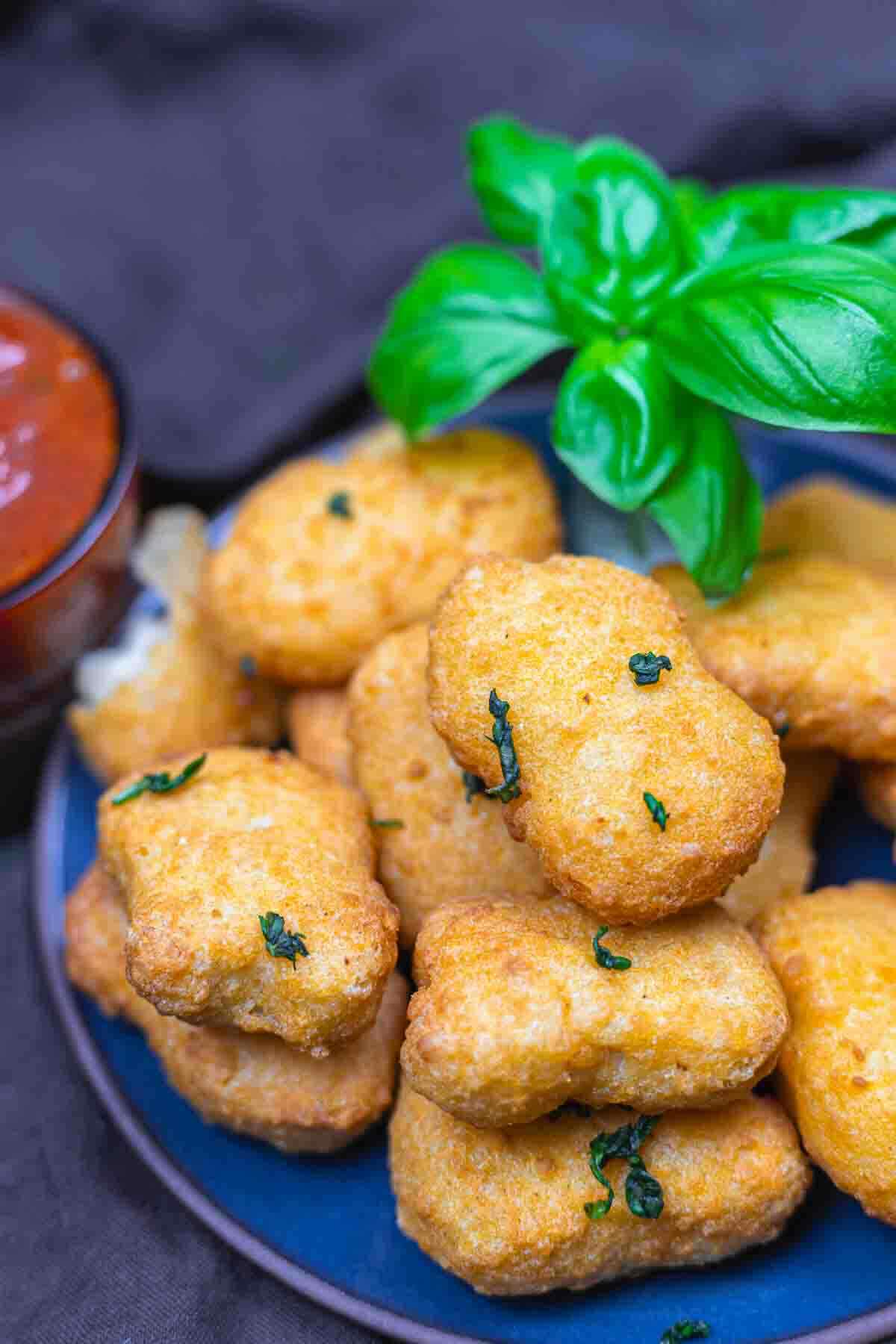 A plate of golden-brown nuggets garnished with chopped herbs, with a fresh basil sprig on top and a bowl of red dipping sauce in the background.