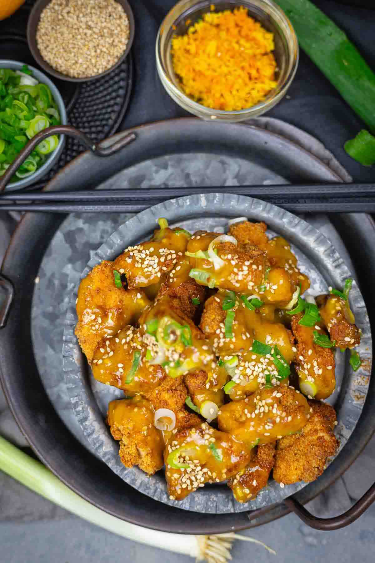 A plate of breaded, sauce-coated cauliflower pieces garnished with sesame seeds and chopped green onions, with bowls of sesame seeds and grated carrot nearby.