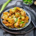 A metal plate with pieces of fried chicken coated in a yellow sauce, garnished with sesame seeds and chopped green onions. Chopsticks and a small bowl of herbs are nearby.
