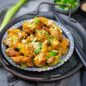A metal plate with pieces of fried chicken coated in a yellow sauce, garnished with sesame seeds and chopped green onions. Chopsticks and a small bowl of herbs are nearby.