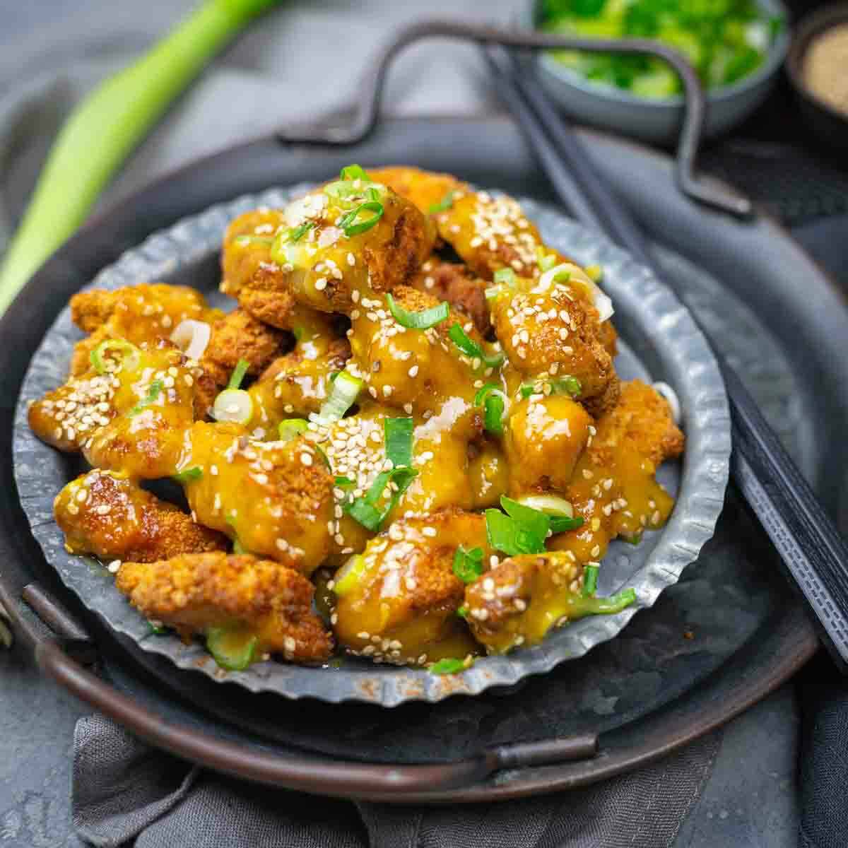 A metal plate with pieces of fried chicken coated in a yellow sauce, garnished with sesame seeds and chopped green onions. Chopsticks and a small bowl of herbs are nearby.