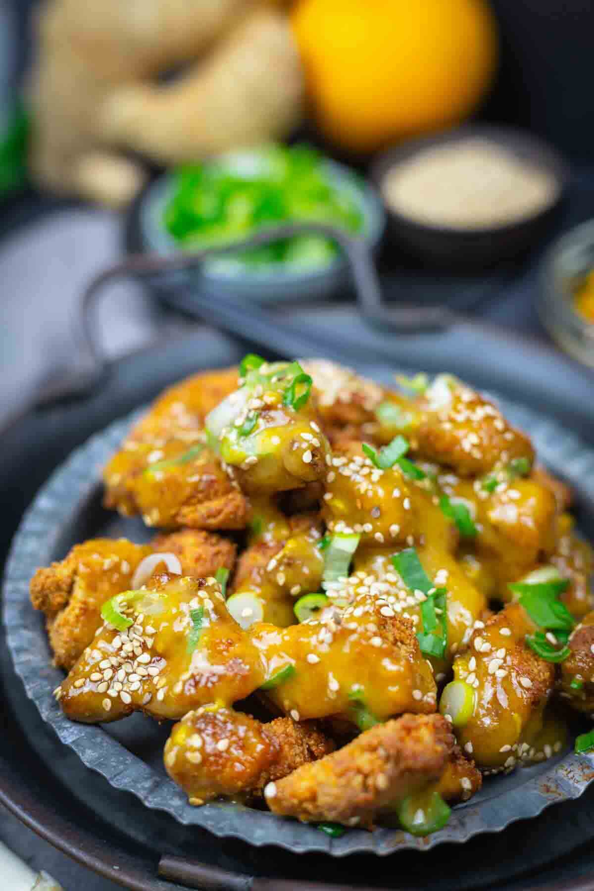 A plate of crispy chicken pieces topped with a golden sauce, garnished with sesame seeds and chopped green onions.