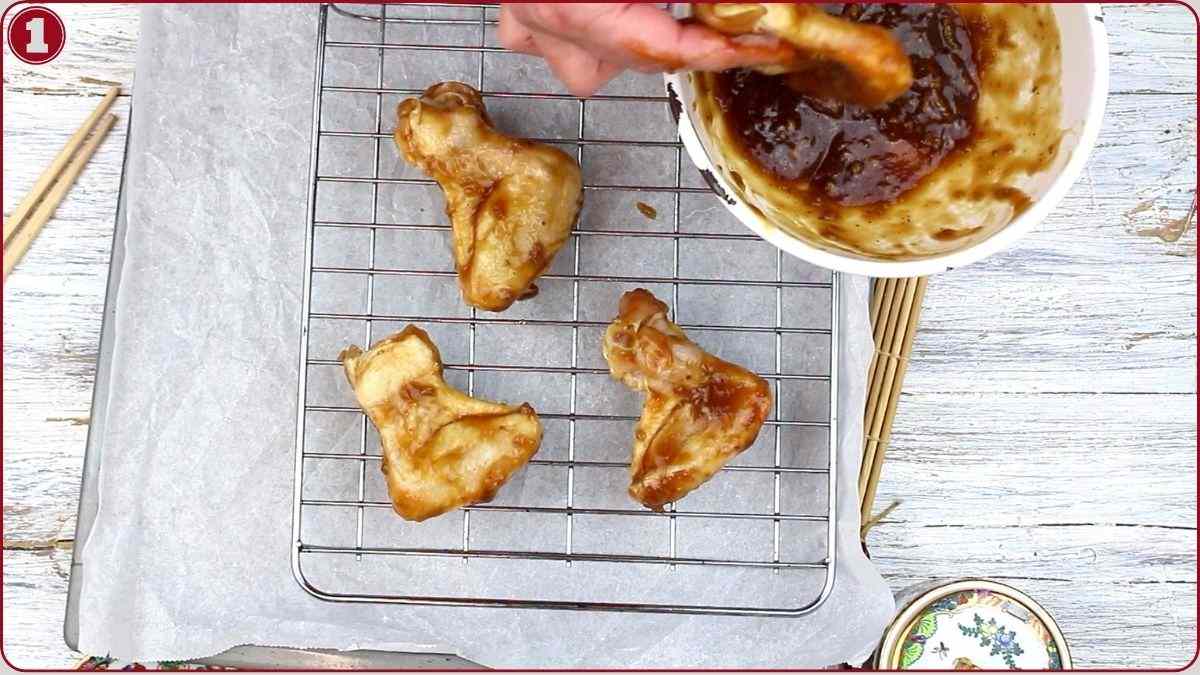 A hand dips a piece of cooked chicken wing into sauce above a bowl, with three sauced wings resting on a wire rack over parchment paper.