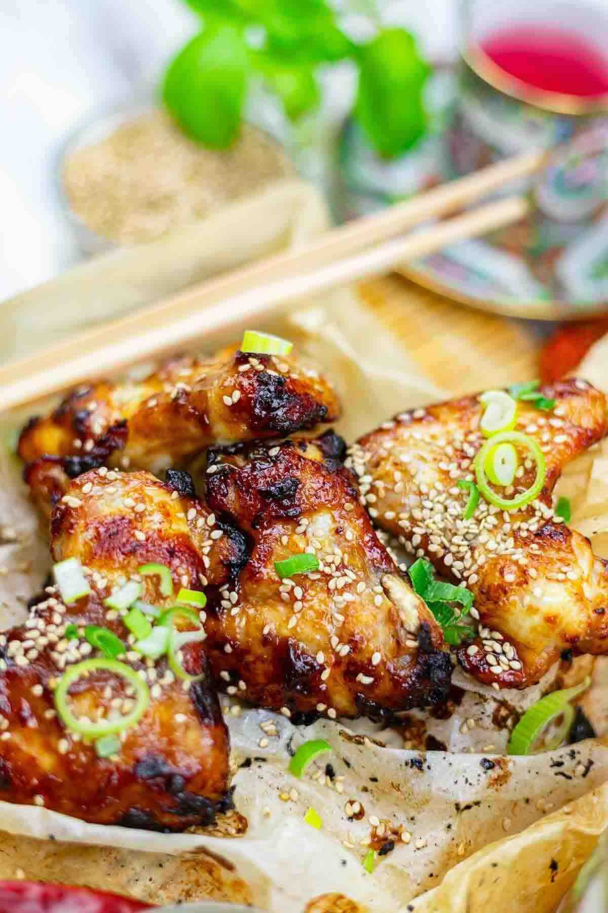 Grilled chicken wings topped with sesame seeds and sliced green onions, served on parchment paper with chopsticks nearby.