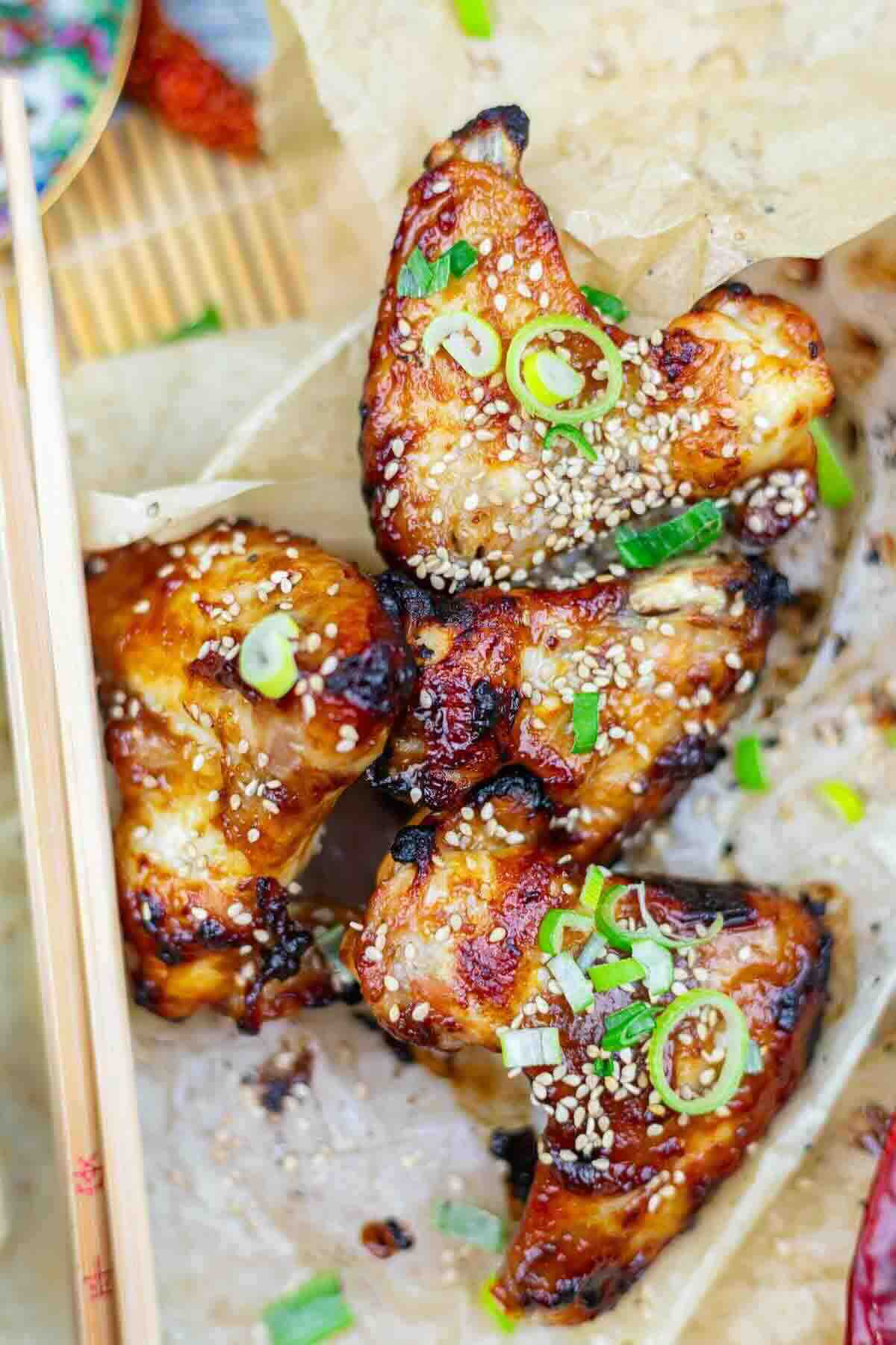 Grilled chicken wings coated in a glossy sauce, garnished with sesame seeds and chopped green onions, served on parchment paper with wooden chopsticks nearby.
