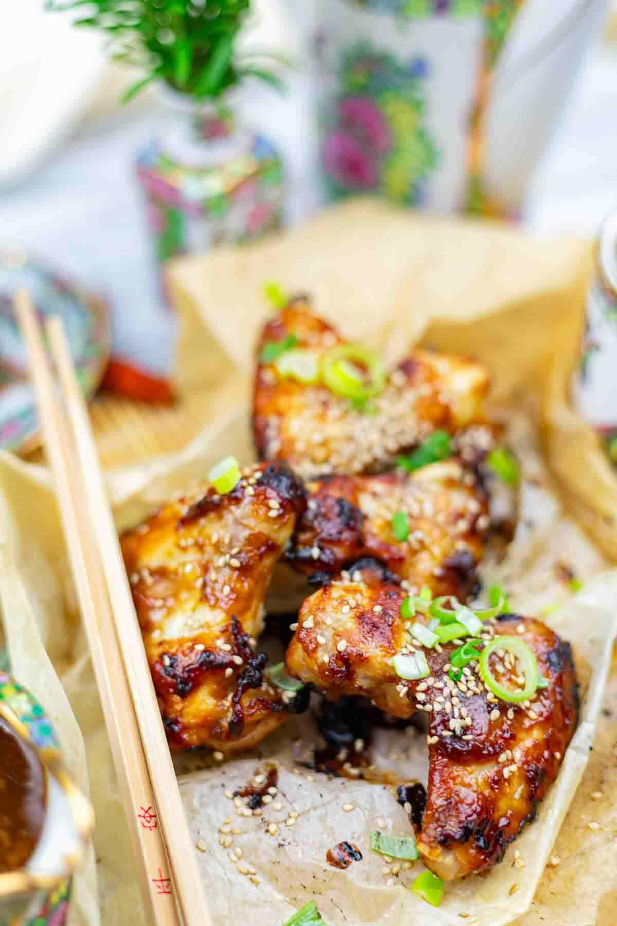 Four glazed chicken wings topped with sesame seeds and chopped green onions on parchment paper, with wooden chopsticks placed alongside.