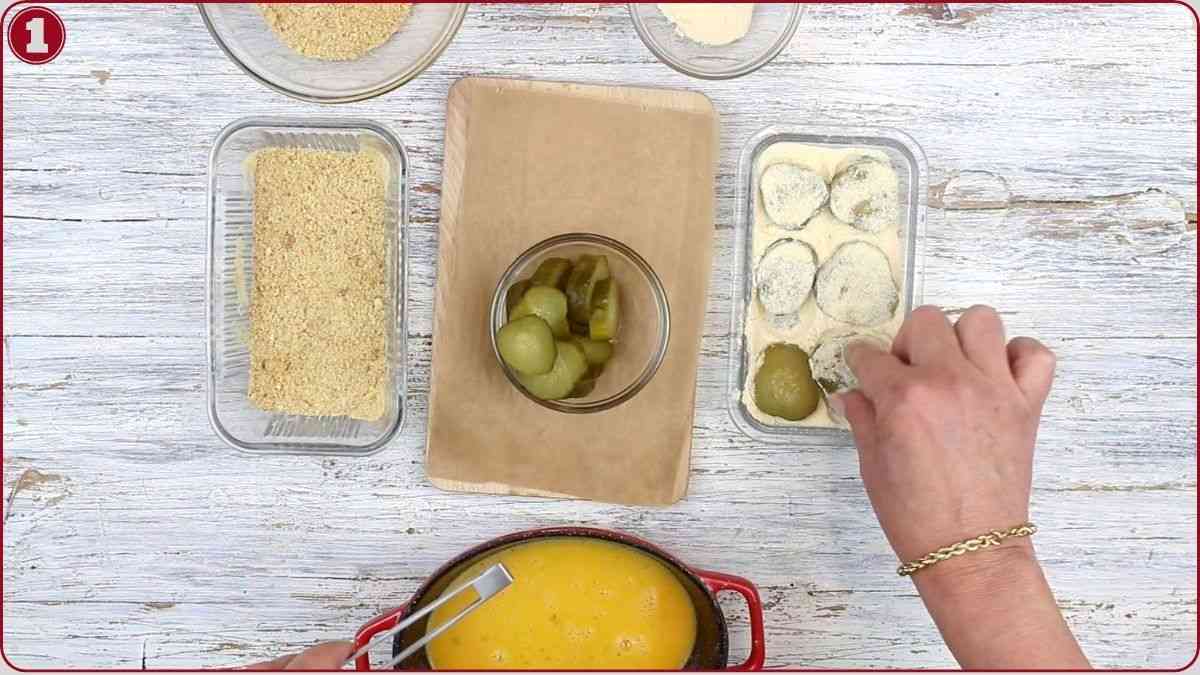 A person dips green olives in batter next to trays of breadcrumbs and beaten eggs on a white wooden surface.