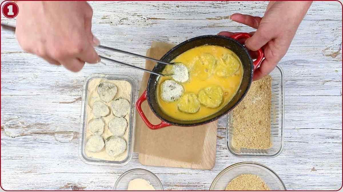 Hands dip breaded zucchini slices into a bowl of beaten eggs with tongs; plates with flour, breadcrumbs, and sliced zucchini are nearby on a wooden surface.
