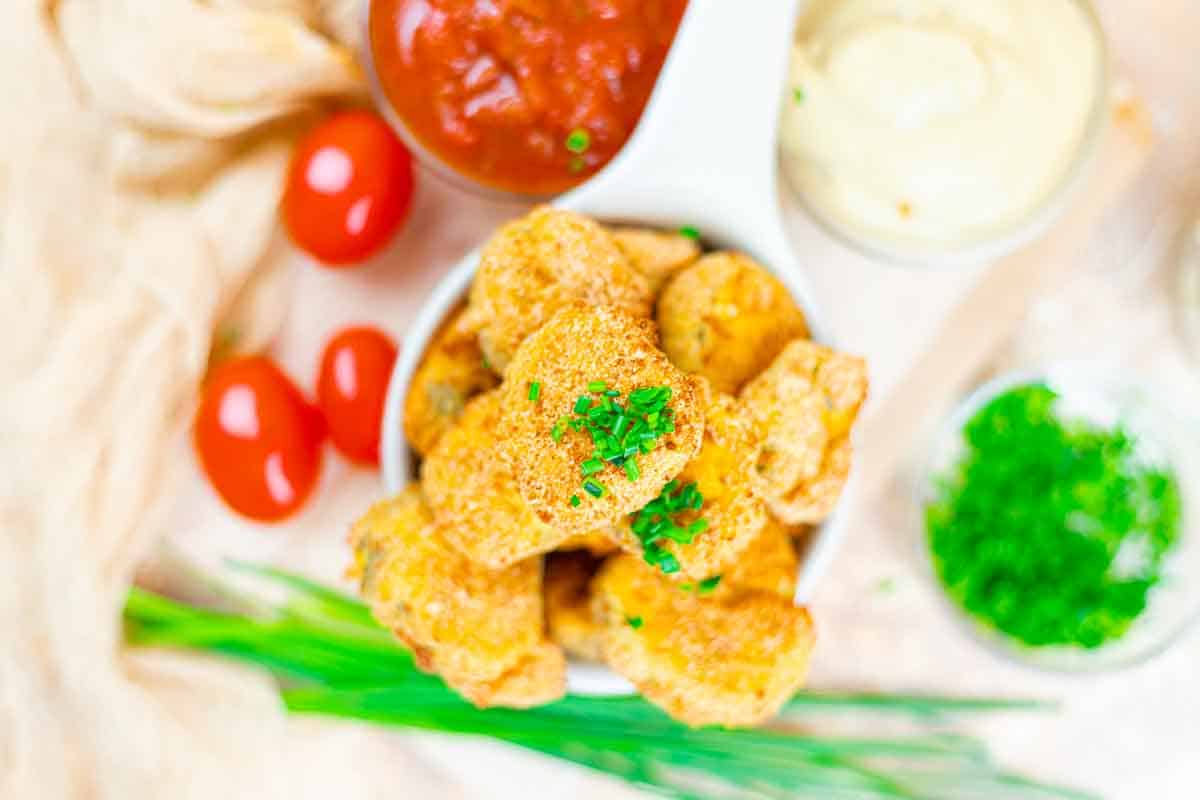 A bowl of breaded pickles garnished with chopped chives, served with red and white dipping sauces, cherry tomatoes, and a small bowl of green herbs.