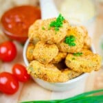 A bowl of golden-brown breaded nuggets topped with chopped chives, surrounded by cherry tomatoes and dipping sauces on a wooden surface.