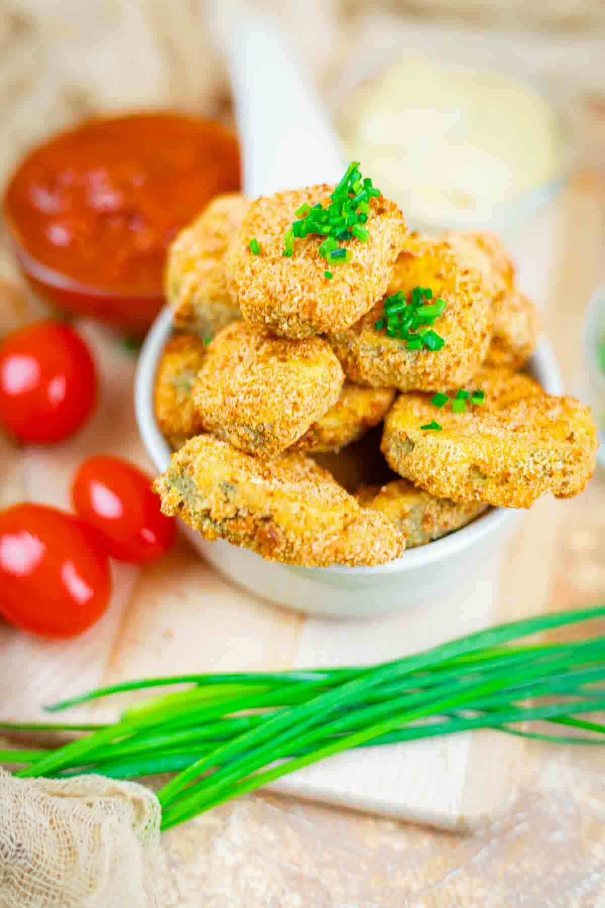 A bowl of breaded, baked zucchini chips garnished with chopped chives, with dipping sauces, cherry tomatoes, and fresh chives in the background.