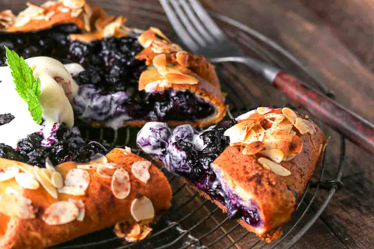 A blueberry galette topped with sliced almonds and a scoop of ice cream sits on a wire rack with a fork beside it.