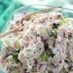 Close-up of a bowl of chicken salad with chopped celery, red onion, and herbs, with a serving spoon in the background.