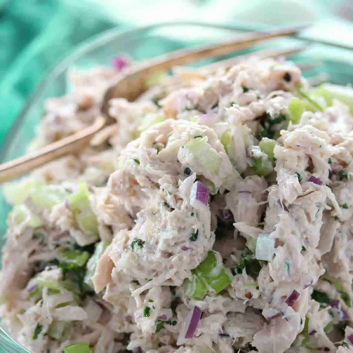 Close-up of a bowl of chicken salad with chopped celery, red onion, and herbs, with a serving spoon in the background.