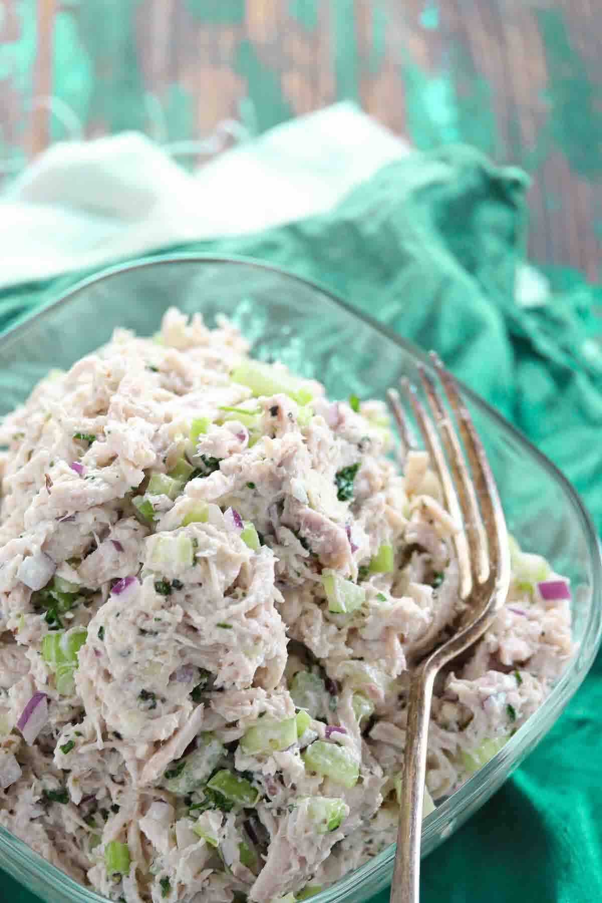 A glass bowl filled with chicken salad containing shredded chicken, chopped celery, and red onion, with a fork resting on the side.