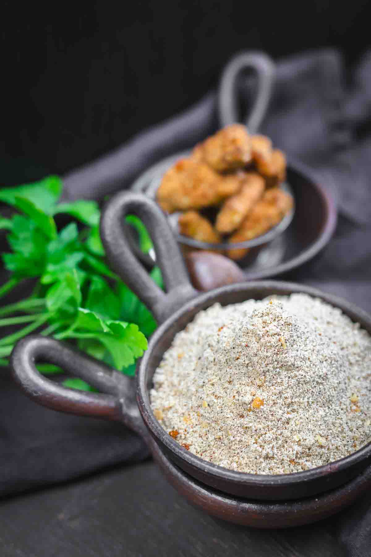 A clay bowl filled with a coarse spice blend sits in the foreground, with fresh herbs and a smaller dish of fried food items in the background.