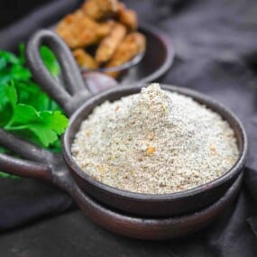 A bowl filled with seasoned flour sits on a dark surface, with herbs and a small dish of fried food in the background.