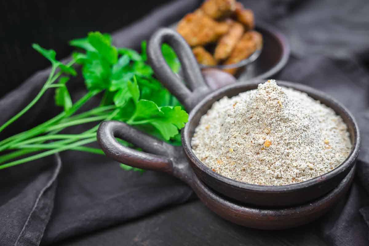 A bowl of chicken breading mix sits on a dark cloth, with fresh parsley and a small dish of fried food in the background.