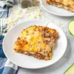 A slice of baked lasagna with ground meat and melted cheese on a white plate, with shredded cheese sprinkled on the table in the background.