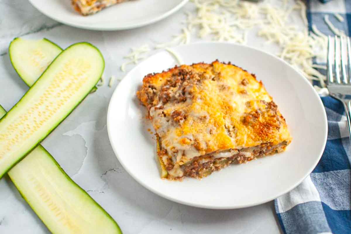 A slice of zucchini lasagna with melted cheese and ground meat is served on a white plate, next to two raw zucchini slices.