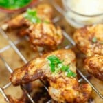 Grilled chicken wings garnished with chopped green herbs, resting on a metal rack with a blurred background of side dishes.