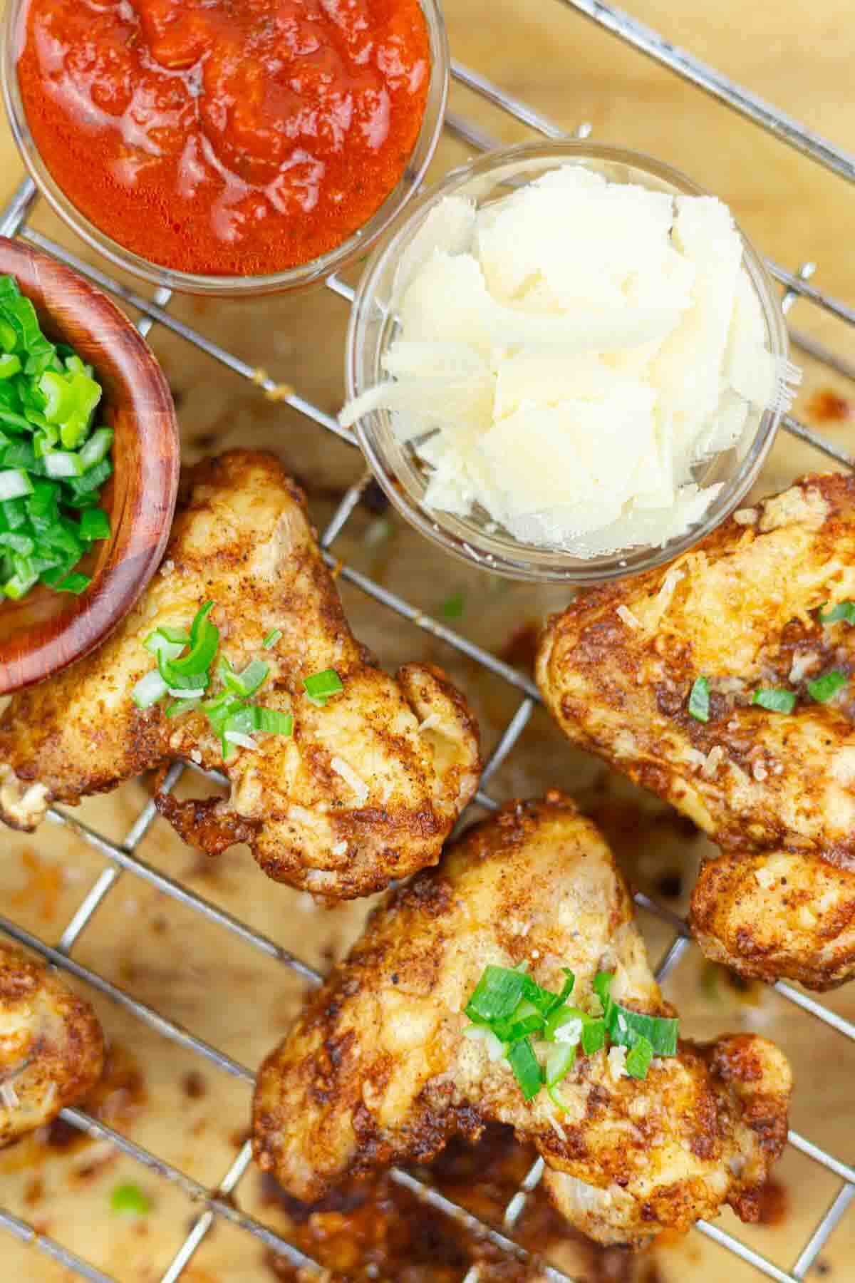 Grilled cauliflower florets on a wire rack, garnished with chopped herbs, with small bowls of tomato sauce, grated cheese, and chopped green onions nearby.