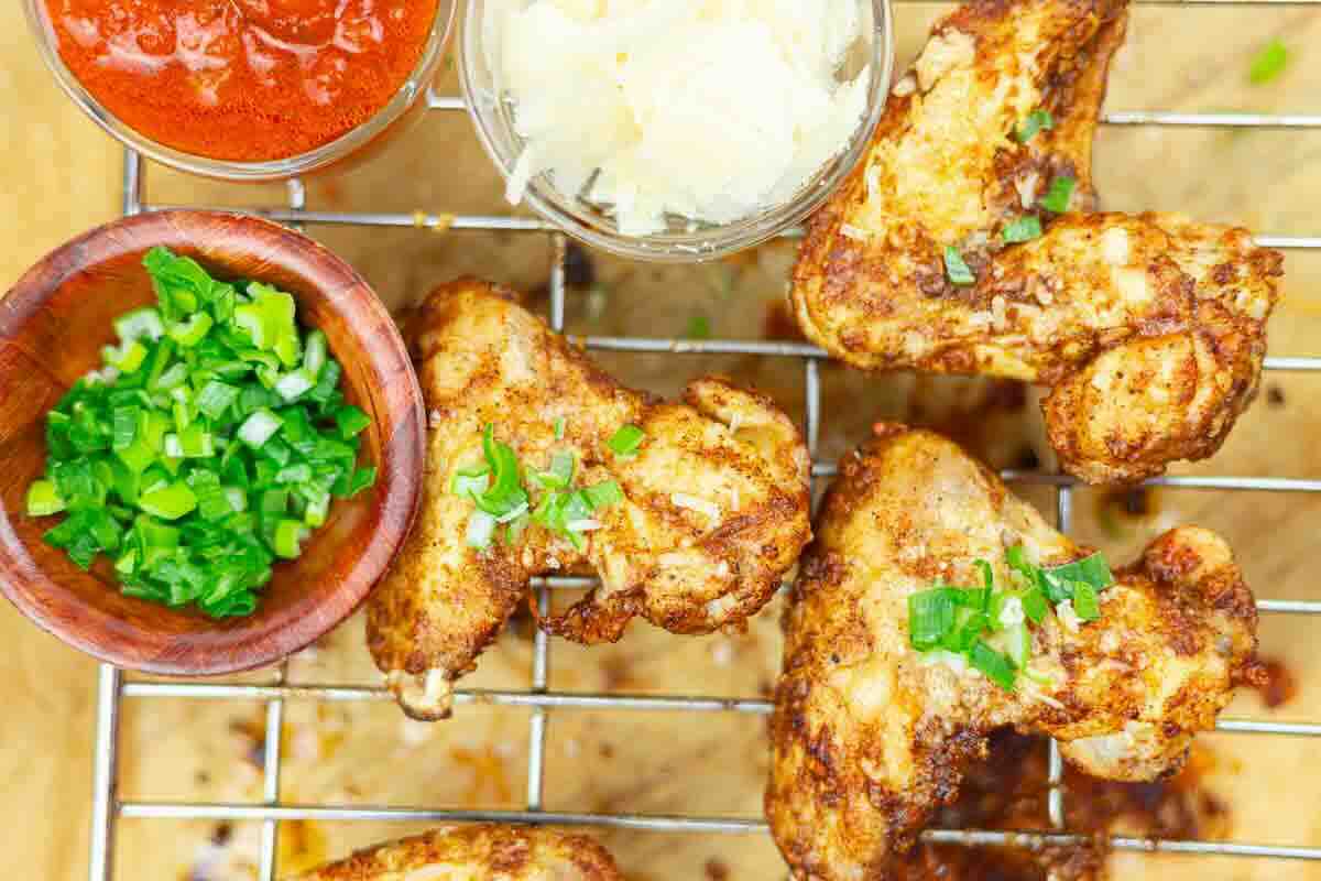 Chicken wings on a wire rack, garnished with chopped green onions, served with bowls of tomato sauce, grated cheese, and more green onions.