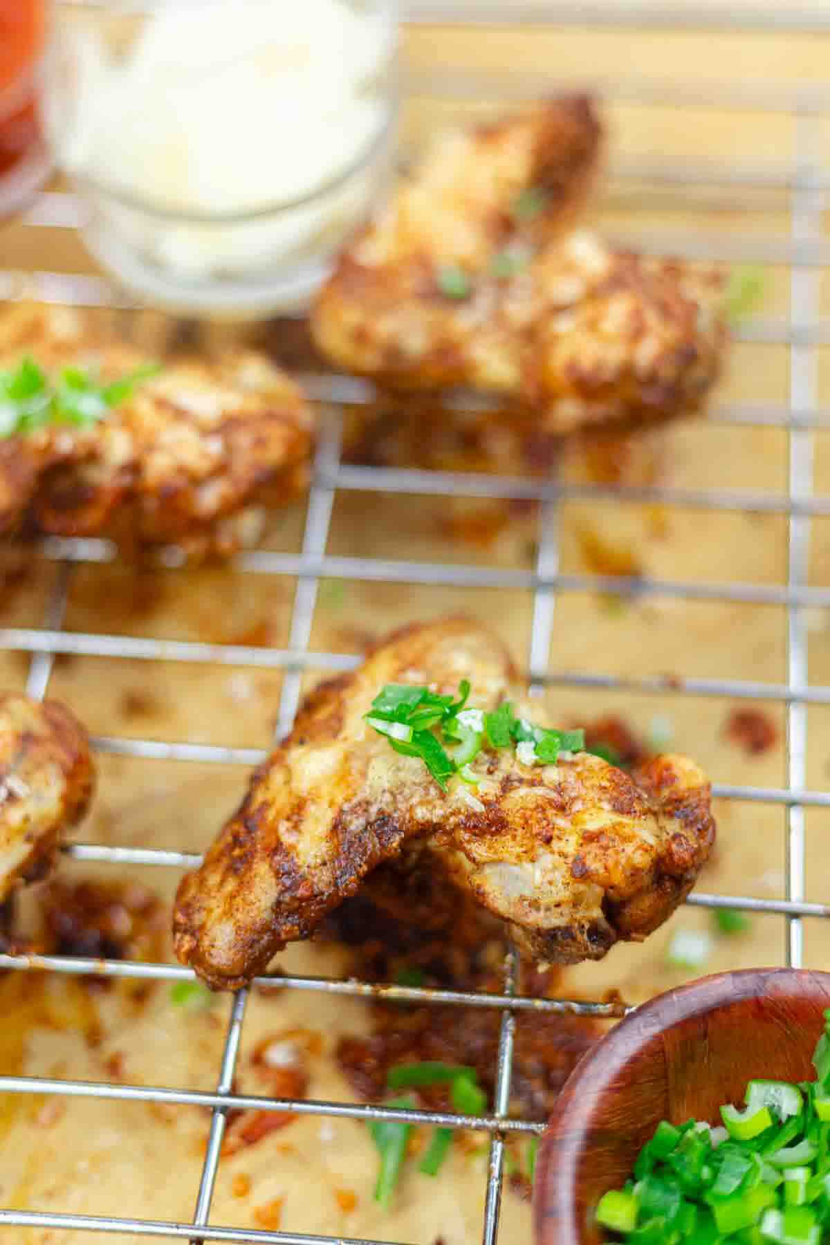 Close-up of cooked chicken wings on a wire rack, garnished with chopped green herbs, with a small bowl of sauce and other food items in the background.