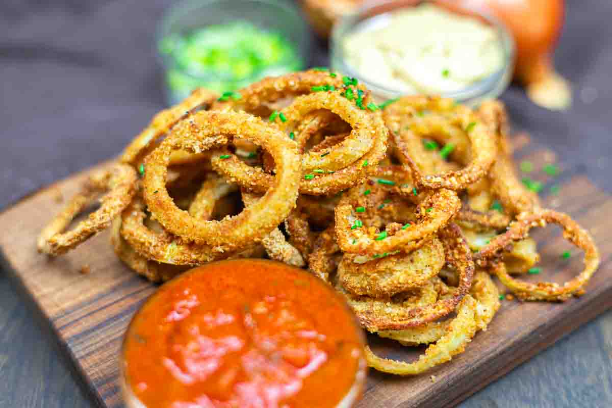 A pile of crispy fried onion rings on a wooden board, garnished with chopped herbs, served with red sauce and a creamy dip in small bowls.