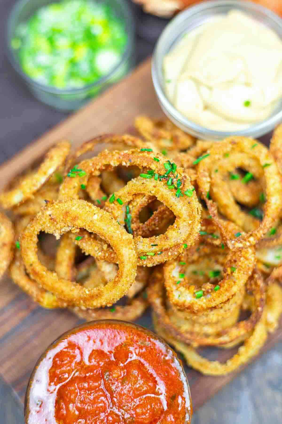 A close-up of crispy onion rings garnished with herbs on a wooden board, served with small bowls of ketchup, dip, and chopped green onions.