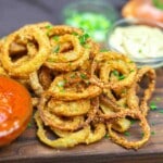 A pile of crispy fried onion rings garnished with chopped chives, served on a wooden board with red and yellow dipping sauces in small bowls.