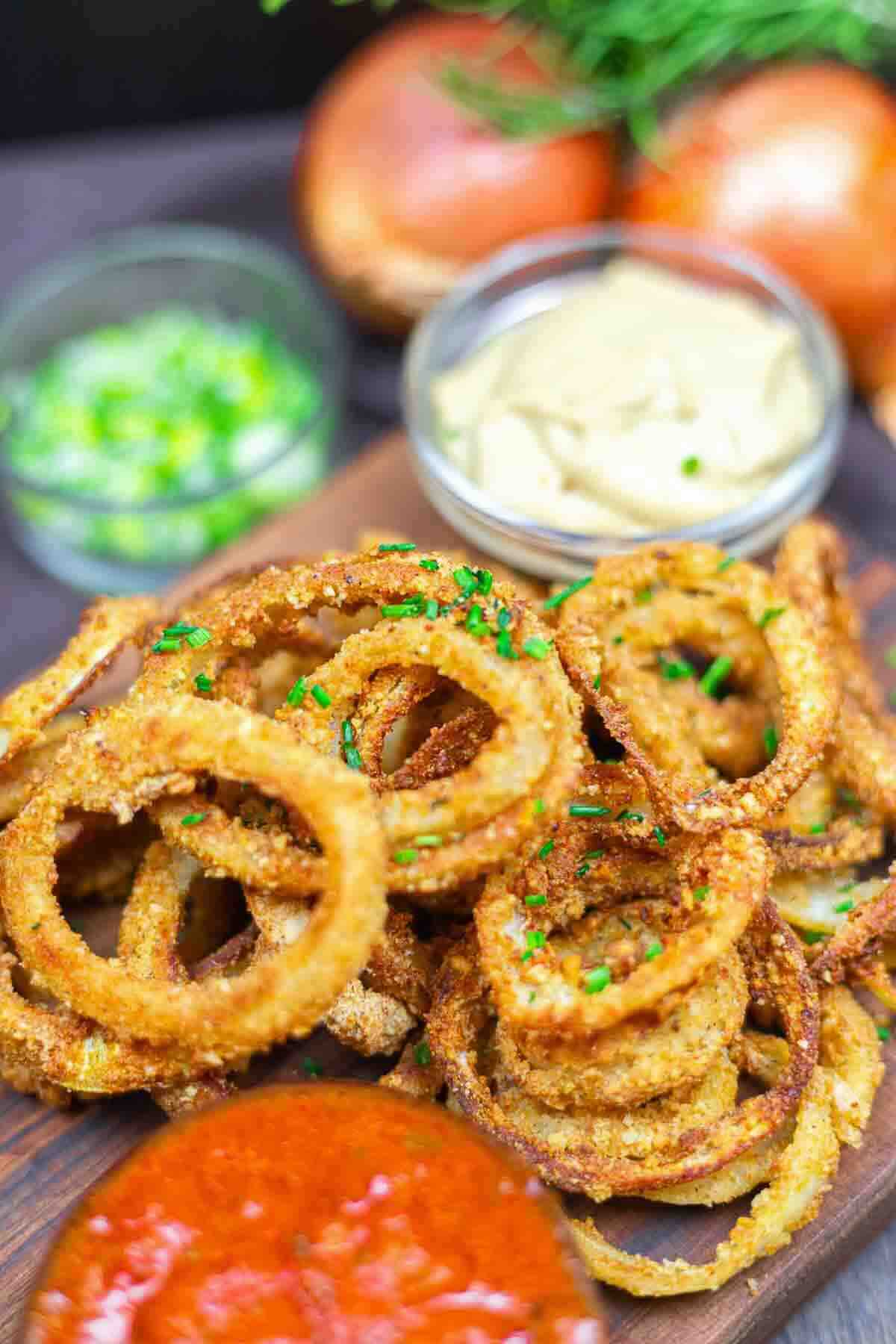 A plate of crispy onion rings garnished with chopped chives, served with small bowls of dipping sauces and fresh vegetables in the background.