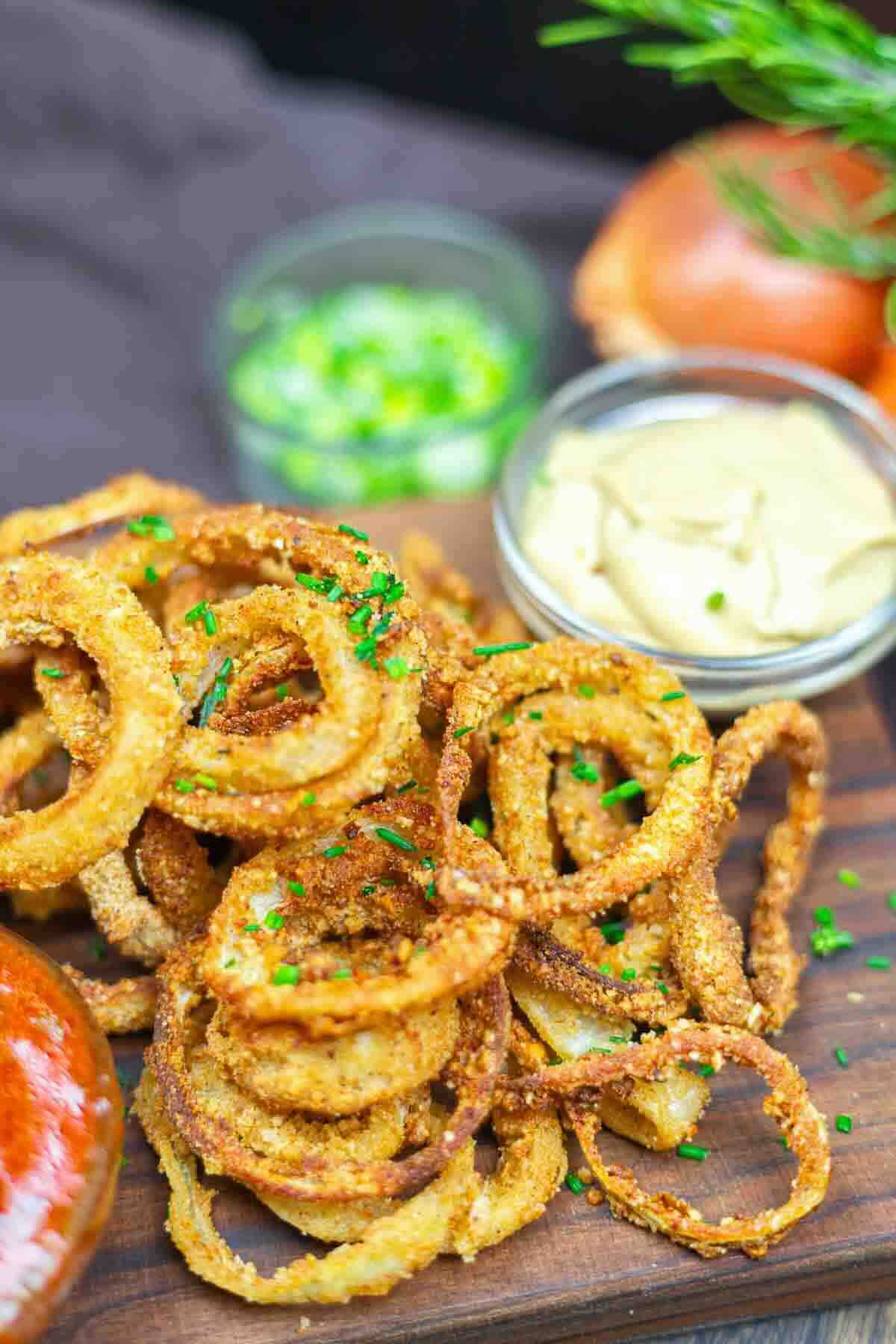 A pile of crispy fried onion rings garnished with chopped chives on a wooden board, served with dipping sauce and chopped green onions in small bowls.