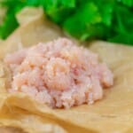 Raw ground chicken placed on brown parchment paper, with green leafy herbs in the blurred background.