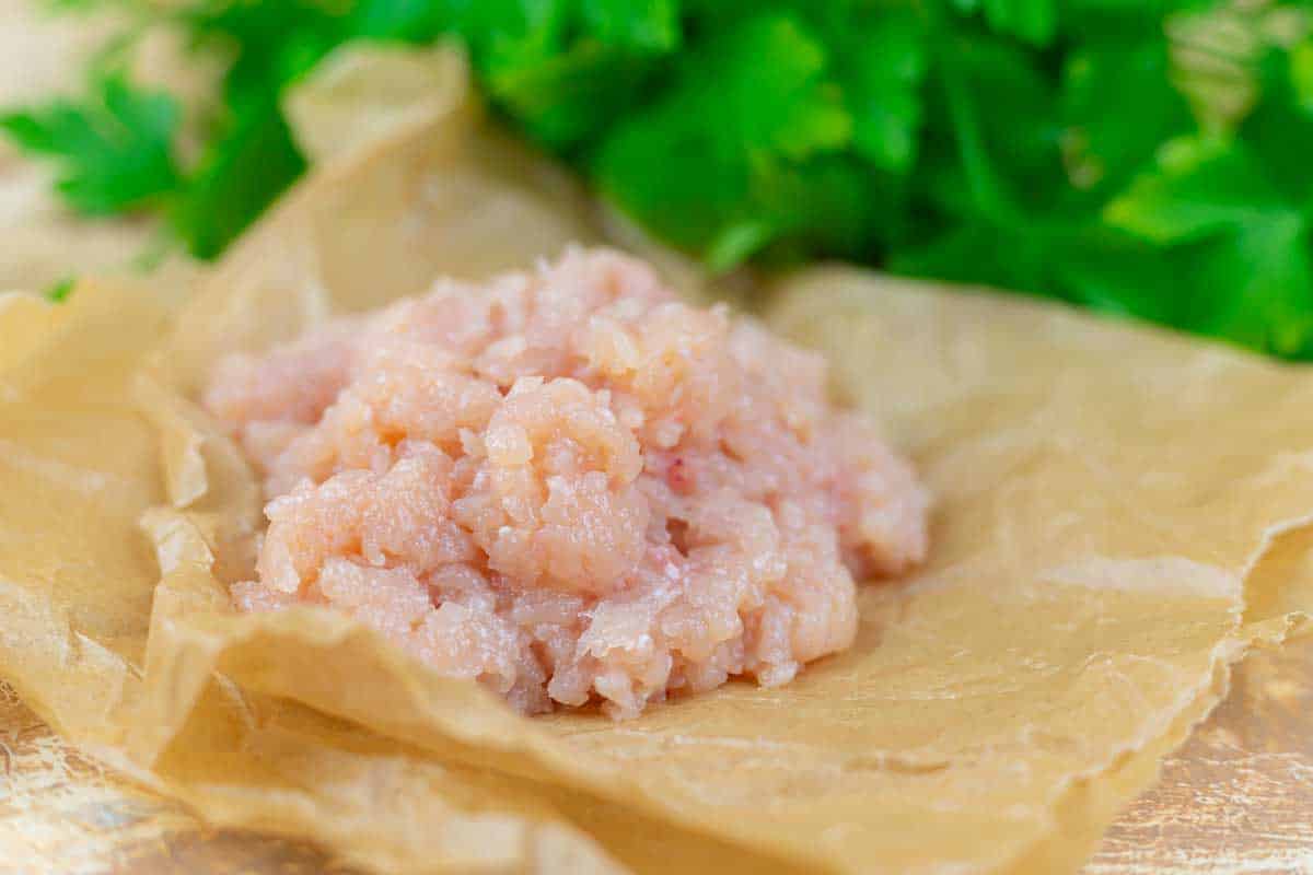 A mound of raw ground chicken sits on brown parchment paper, with fresh green herbs in the background.