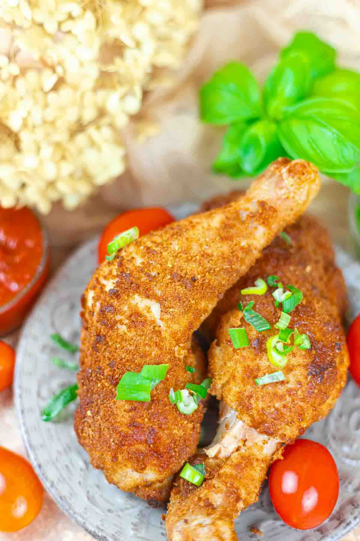 Two breaded and fried chicken drumsticks garnished with chopped green onions, served on a gray plate with cherry tomatoes and fresh basil nearby.