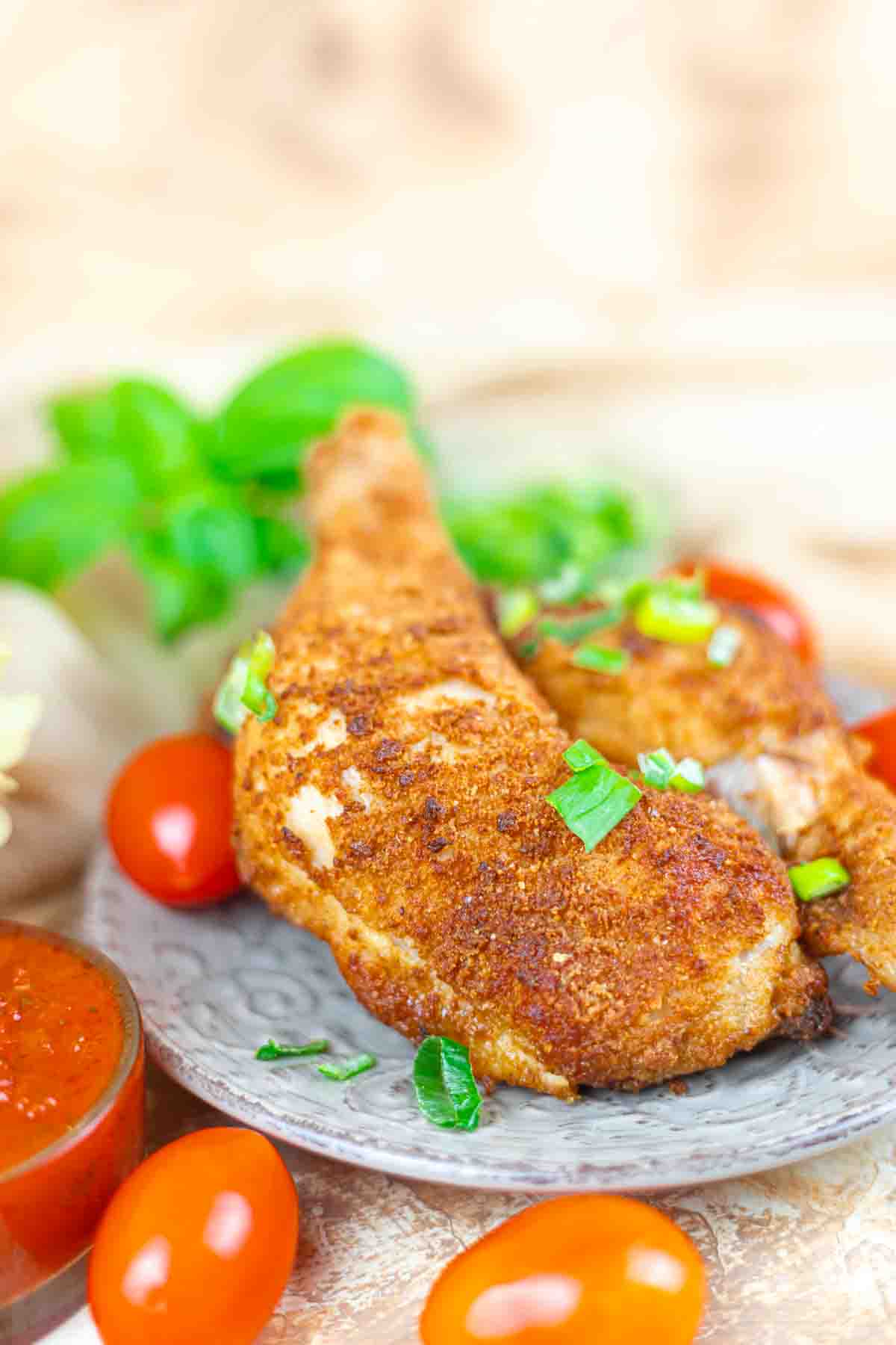 A plate with two breaded and baked chicken legs garnished with chopped herbs, surrounded by cherry tomatoes and a bowl of red sauce.