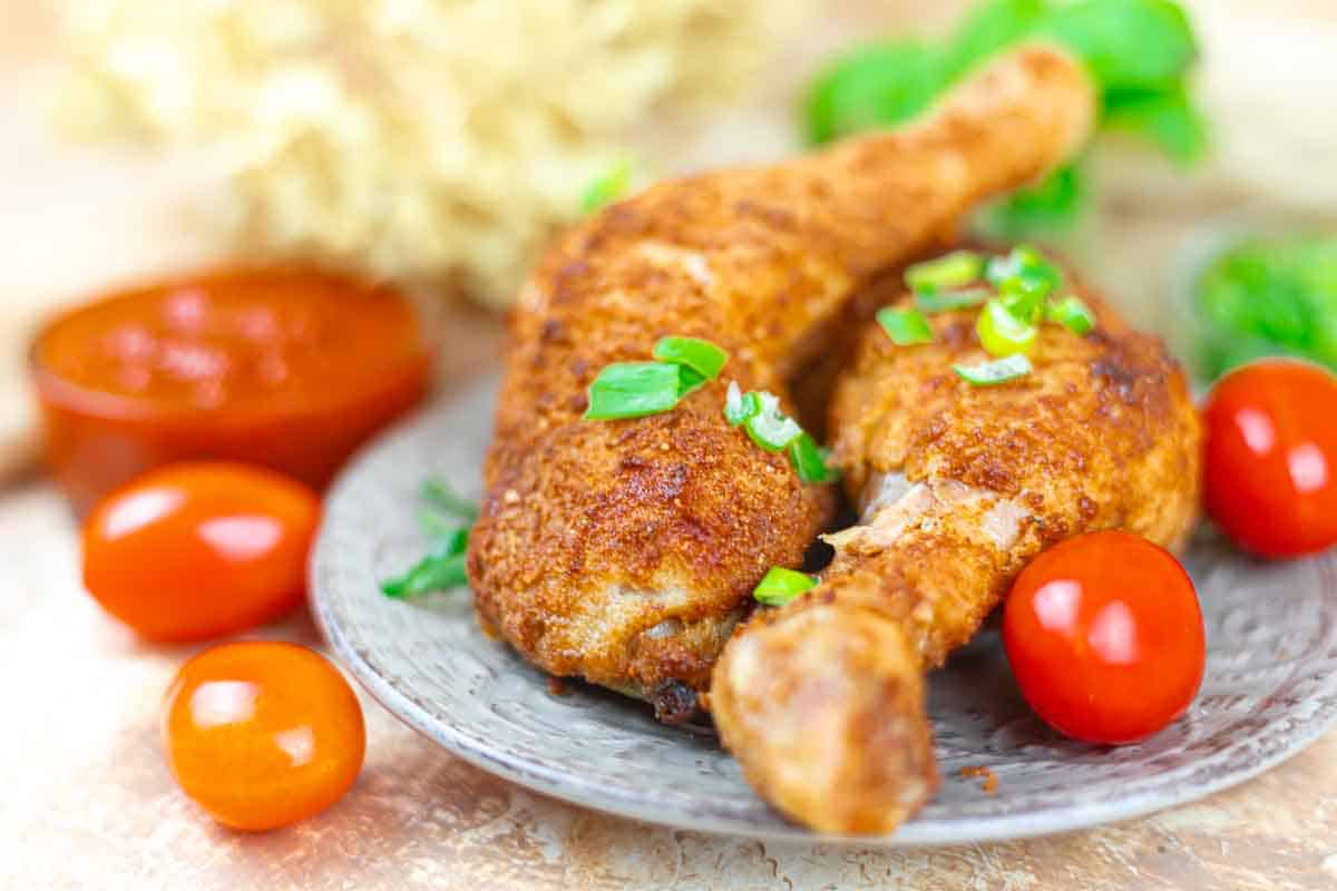 Two seasoned, cooked chicken drumsticks are served on a gray plate with cherry tomatoes and green onion garnish; sauce in a small bowl is visible in the background.