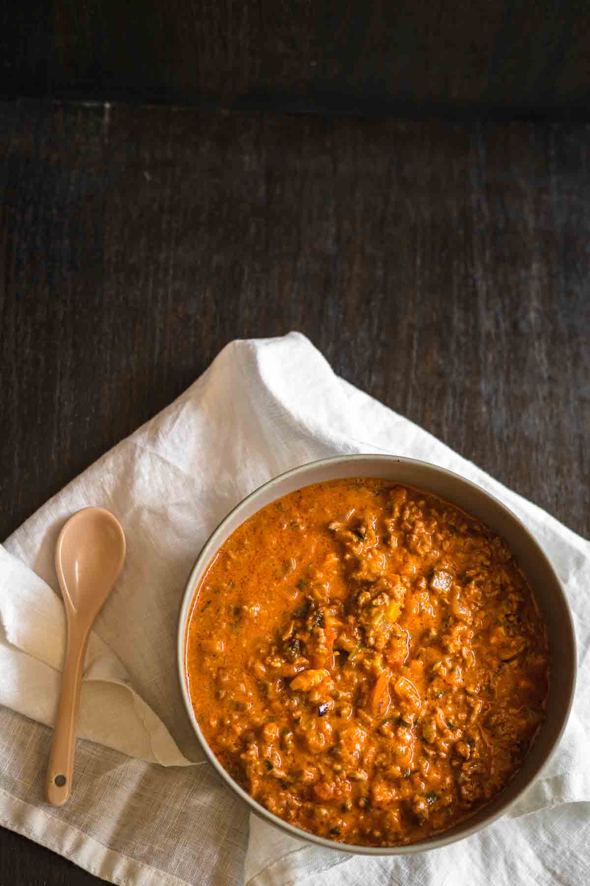 A bowl of tomato-based lentil stew sits on a white cloth napkin next to a beige spoon, atop a dark wooden surface.