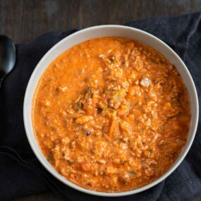 A bowl of thick, orange-colored ground meat stew with visible herbs and vegetables, placed on a dark cloth next to a black spoon.