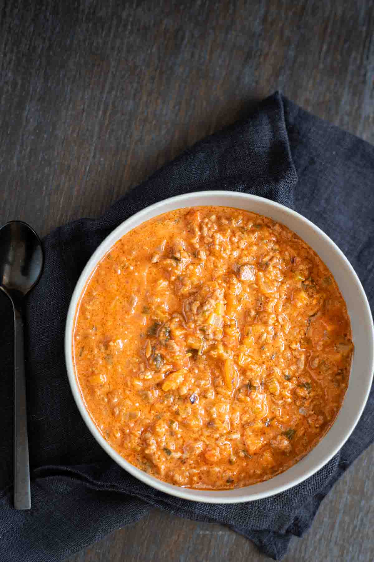 A white bowl filled with creamy tomato-based soup with ground meat and vegetables, placed on a dark cloth next to a black spoon.