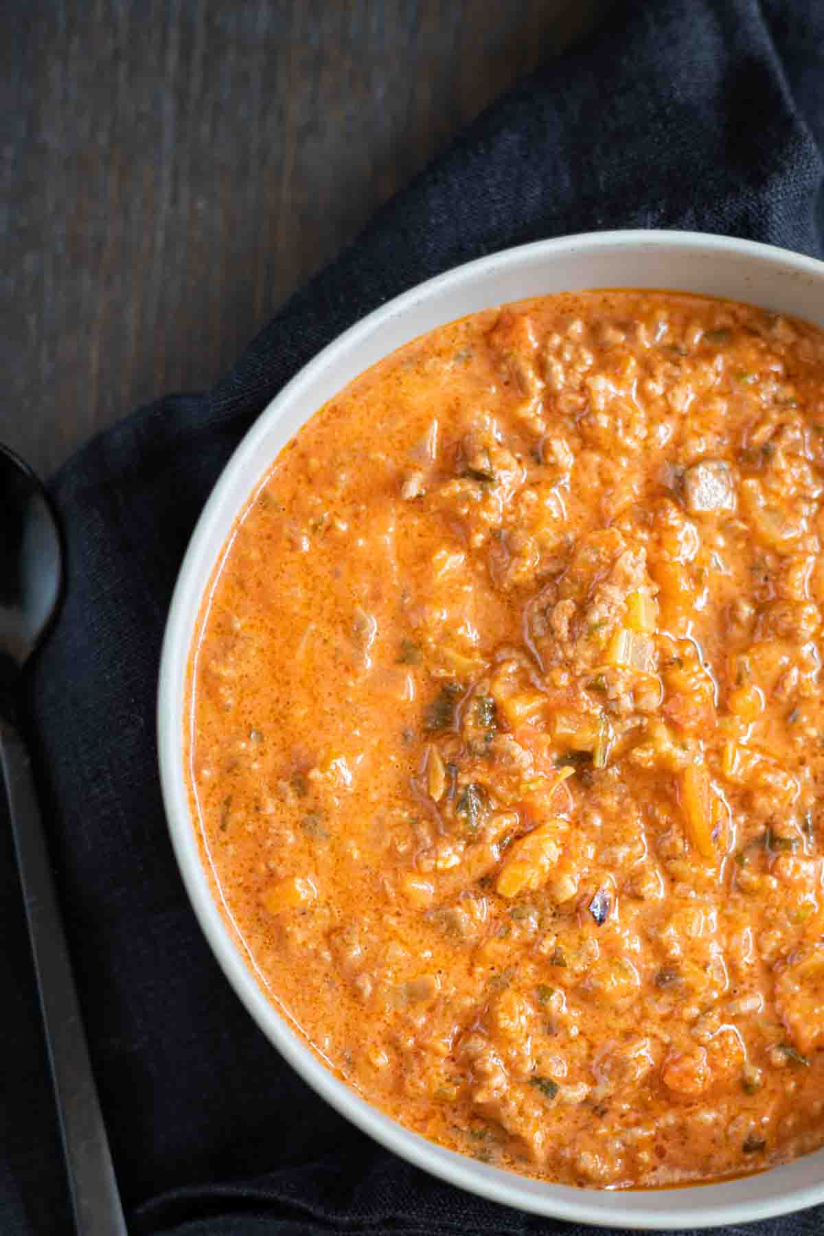 A close-up of a bowl of thick, creamy tomato-based soup with visible chunks of ground meat and vegetables, set on a dark cloth with a black spoon beside it.